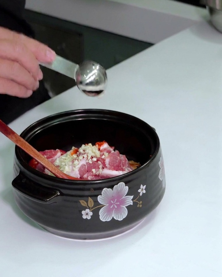 A hand shaking red chili powder from a glass jar onto a wooden spoon held over a pot of ingredients.