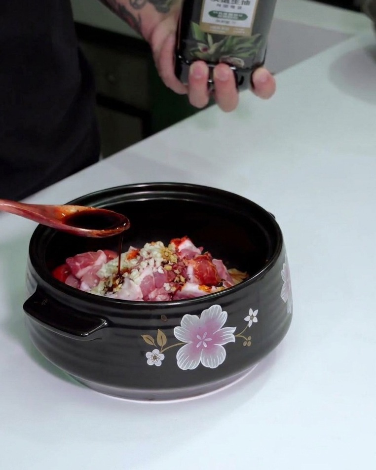 A hand pouring light soy sauce from a bottle into a wooden spoon over the cooking pot.