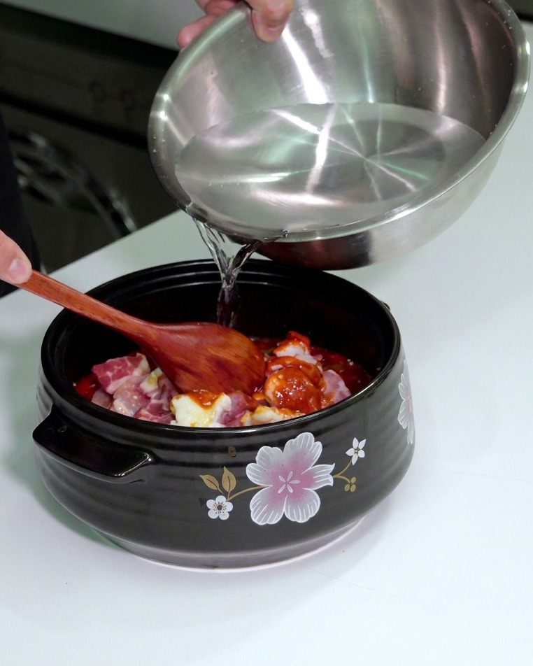 A person pouring water from a stainless steel mixing bowl into a ceramic pot containing seasoned meat and vegetables.