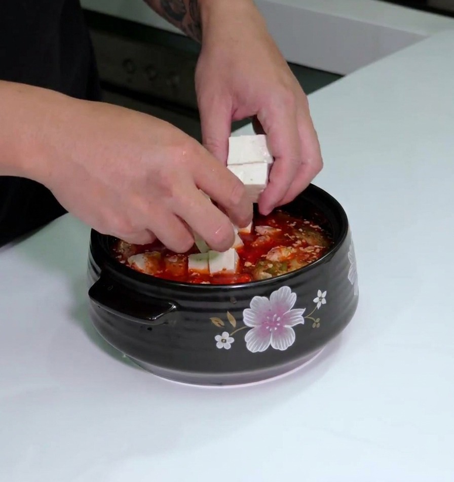 Hands placing fresh white tofu cubes into a black ceramic pot filled with red kimchi soup base and sliced pork belly.