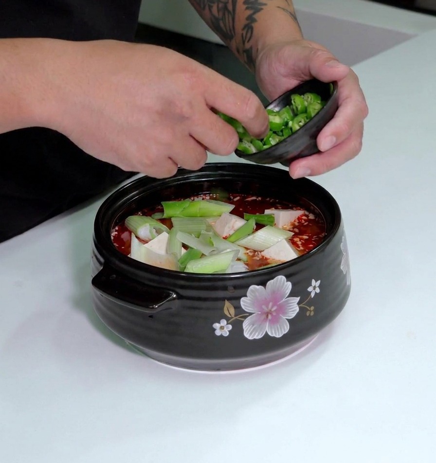 Sliced green chilies being sprinkled from a small bowl onto the top of a pot of kimchi soup containing tofu and leeks.