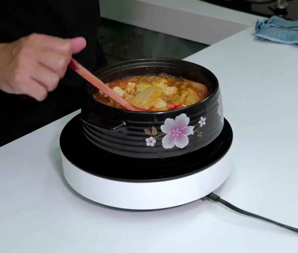 A bottle of Tabasco sauce being held over a steaming, bubbling pot of red kimchi soup while it is stirred with a wooden spoon.