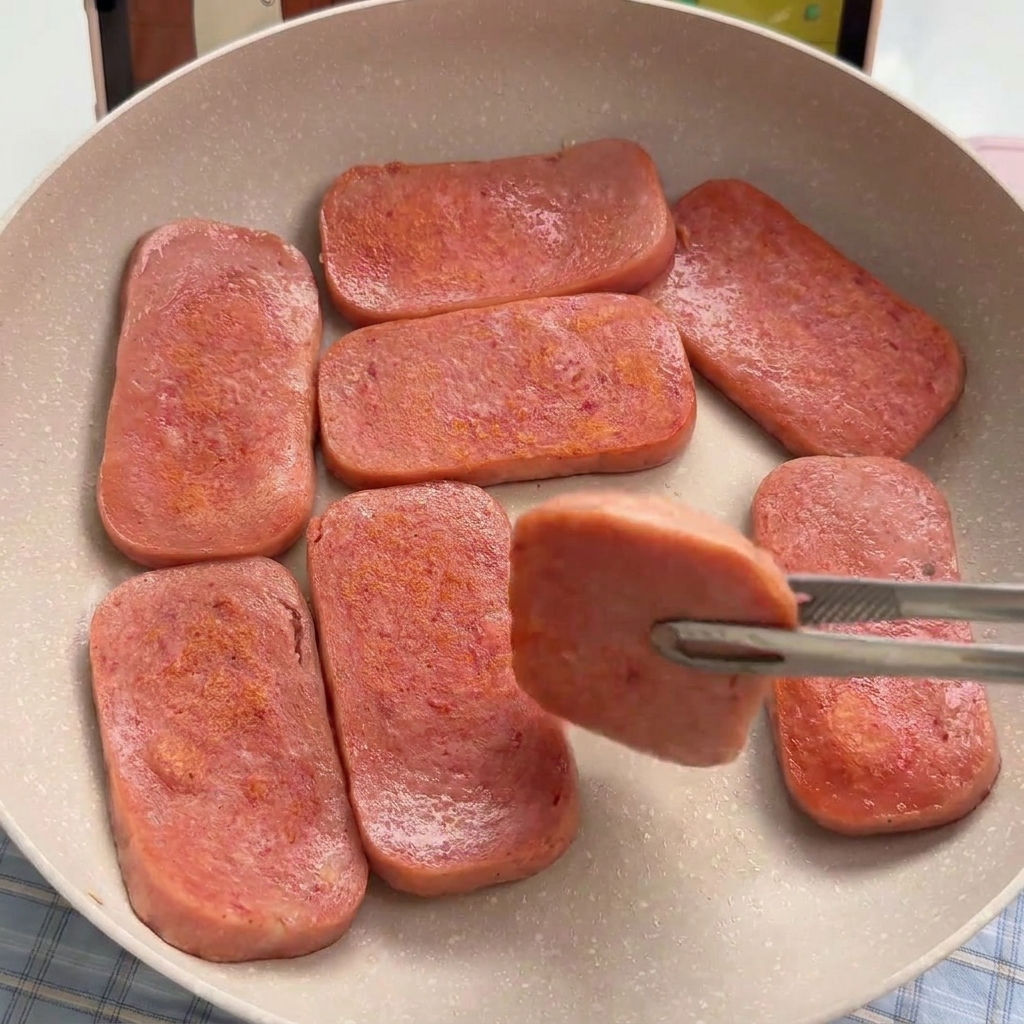 Several rectangular slices of pink luncheon meat being seared in a light-colored non-stick pan until golden brown.