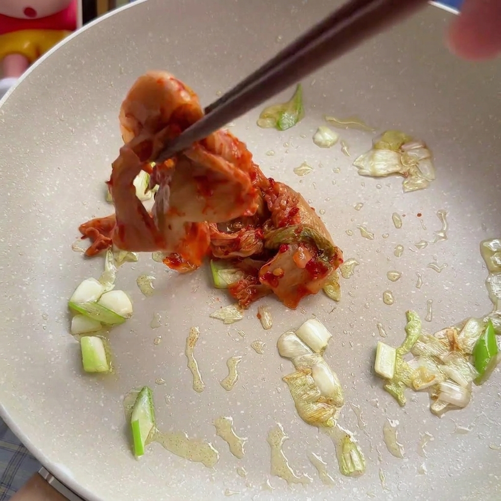 A pair of chopsticks adding a pile of red, chopped cabbage kimchi into a pan with sautéed green onions.