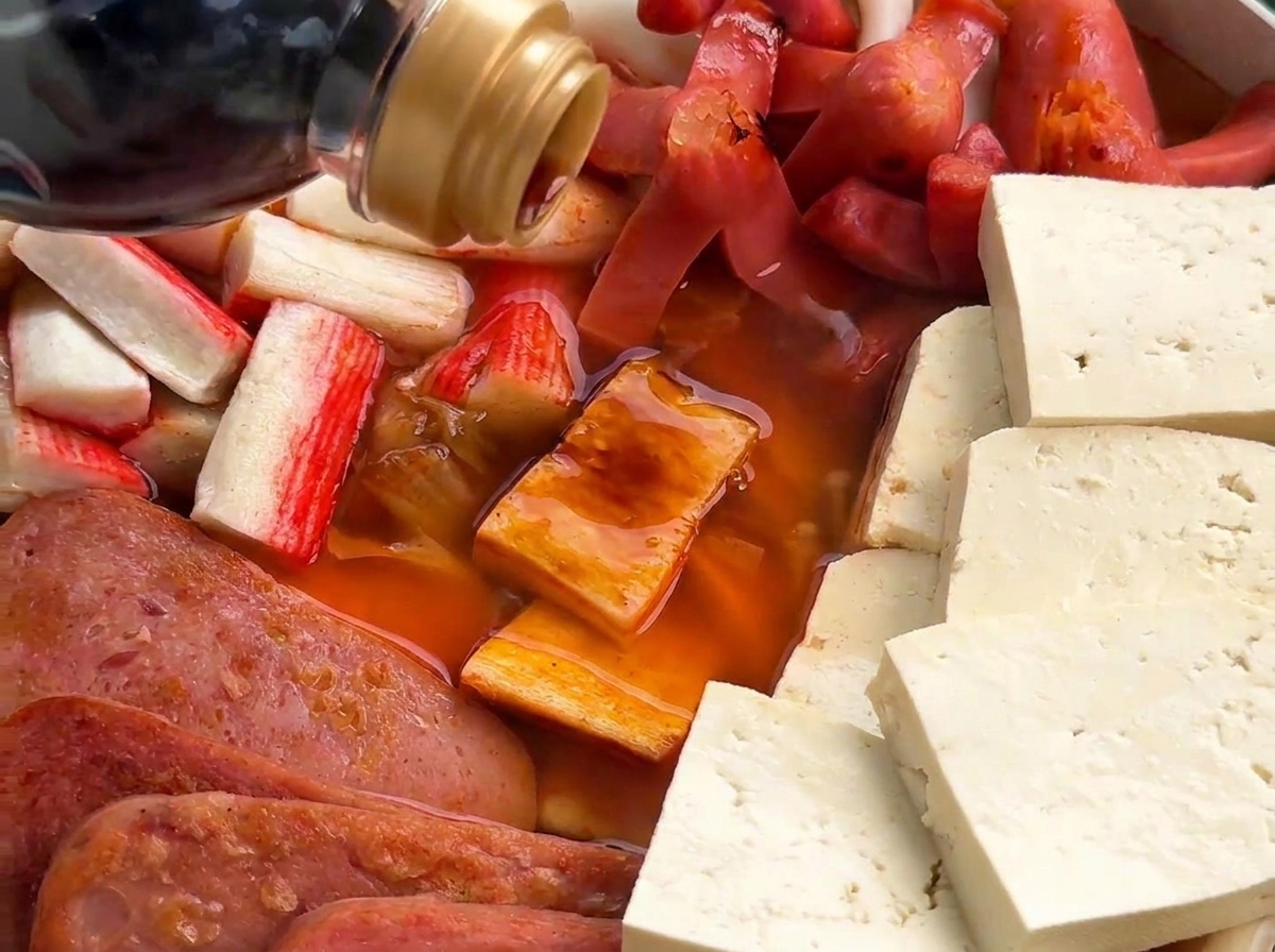 A close-up shot of dark soy sauce being poured from a bottle into a pot filled with sausages, crab sticks, and spam in a reddish-orange broth.