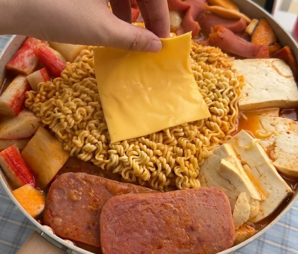 A hand laying a square slice of yellow cheese onto softening ramen noodles in a steaming pot of Korean Army Stew.