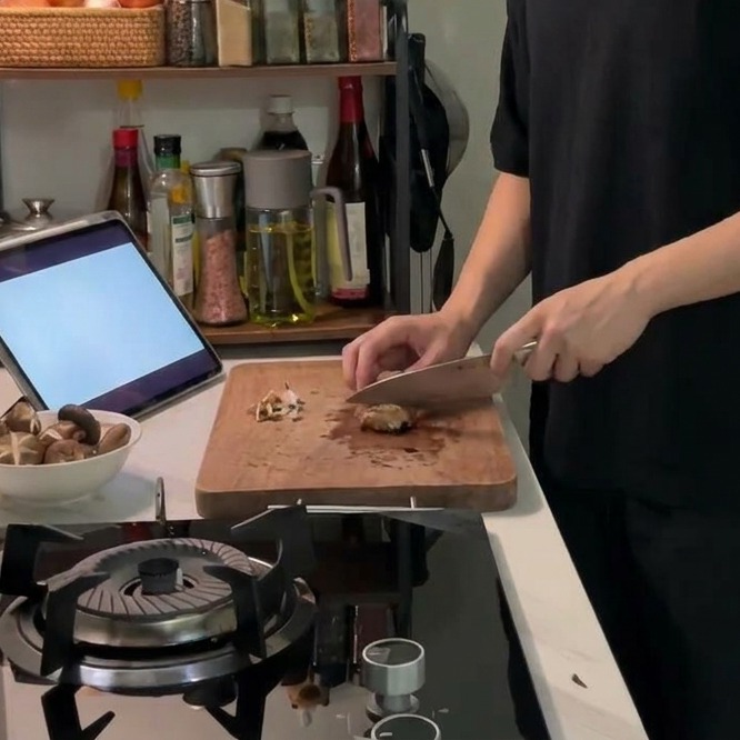 A sharp knife scoring a decorative cross pattern into the top of a fresh brown shiitake mushroom cap on a wooden cutting board.