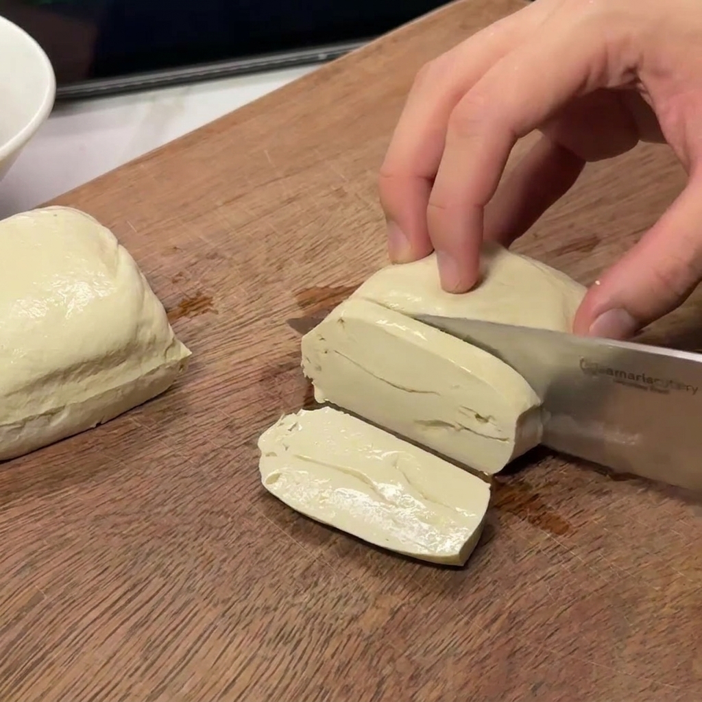 A pair of hands slicing a block of firm white tofu into consistent rectangular pieces on a wooden cutting surface.