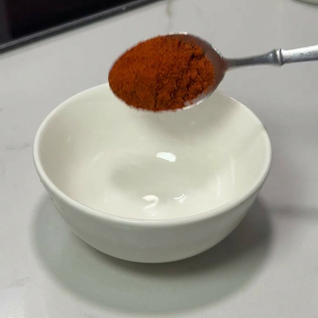 A silver spoon pouring a heap of bright red chili powder into a clean white ceramic bowl.