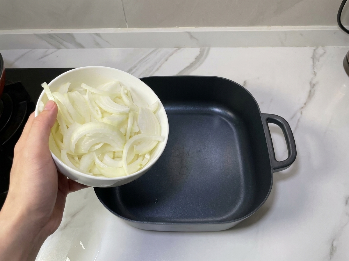 A hand holding a white bowl of sliced onions over an empty black square cooking pot, ready to layer them.