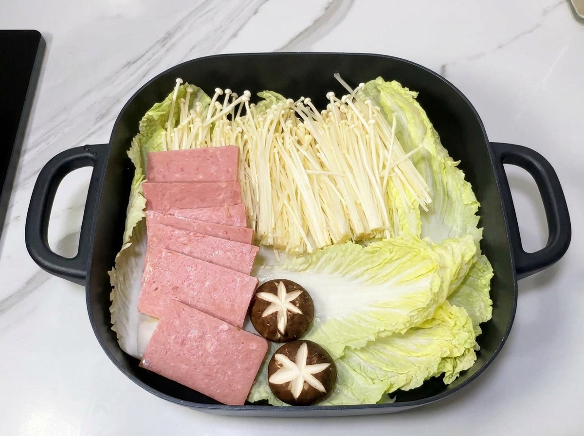 A square pot containing arranged layers of Napa cabbage, enoki mushrooms, and spam slices on a bed of onions.