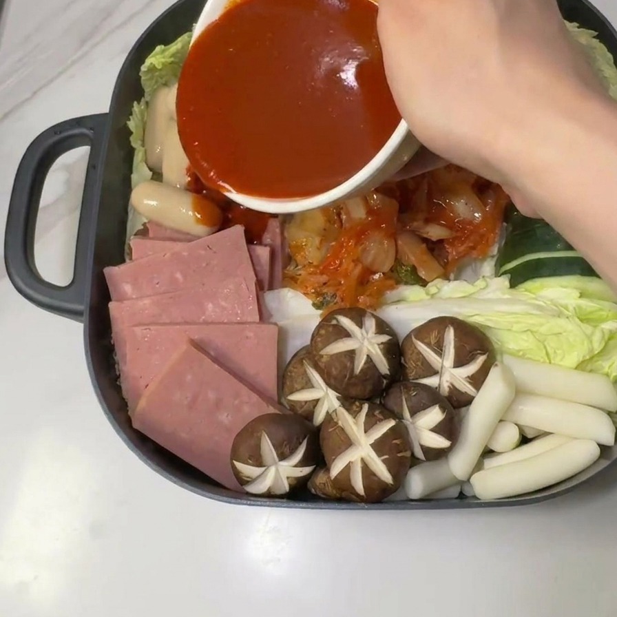 Spicy red gochujang sauce being poured from a white bowl onto the center of the raw army stew ingredients.