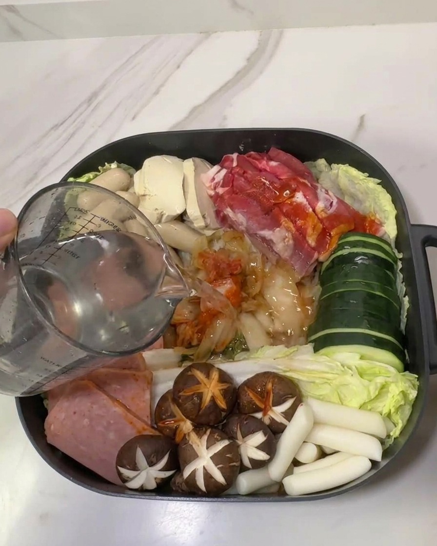 A glass measuring cup pouring water into a pot filled with arranged Korean Army Stew ingredients including spam, mushrooms, and kimchi.