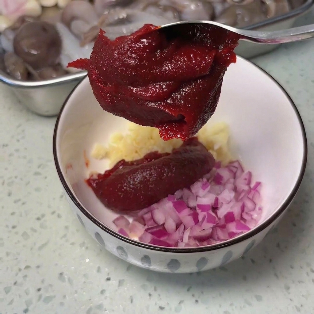 A metal spoon holding a thick dollop of dark red Korean chili paste over a white bowl containing minced garlic and diced red onions.
