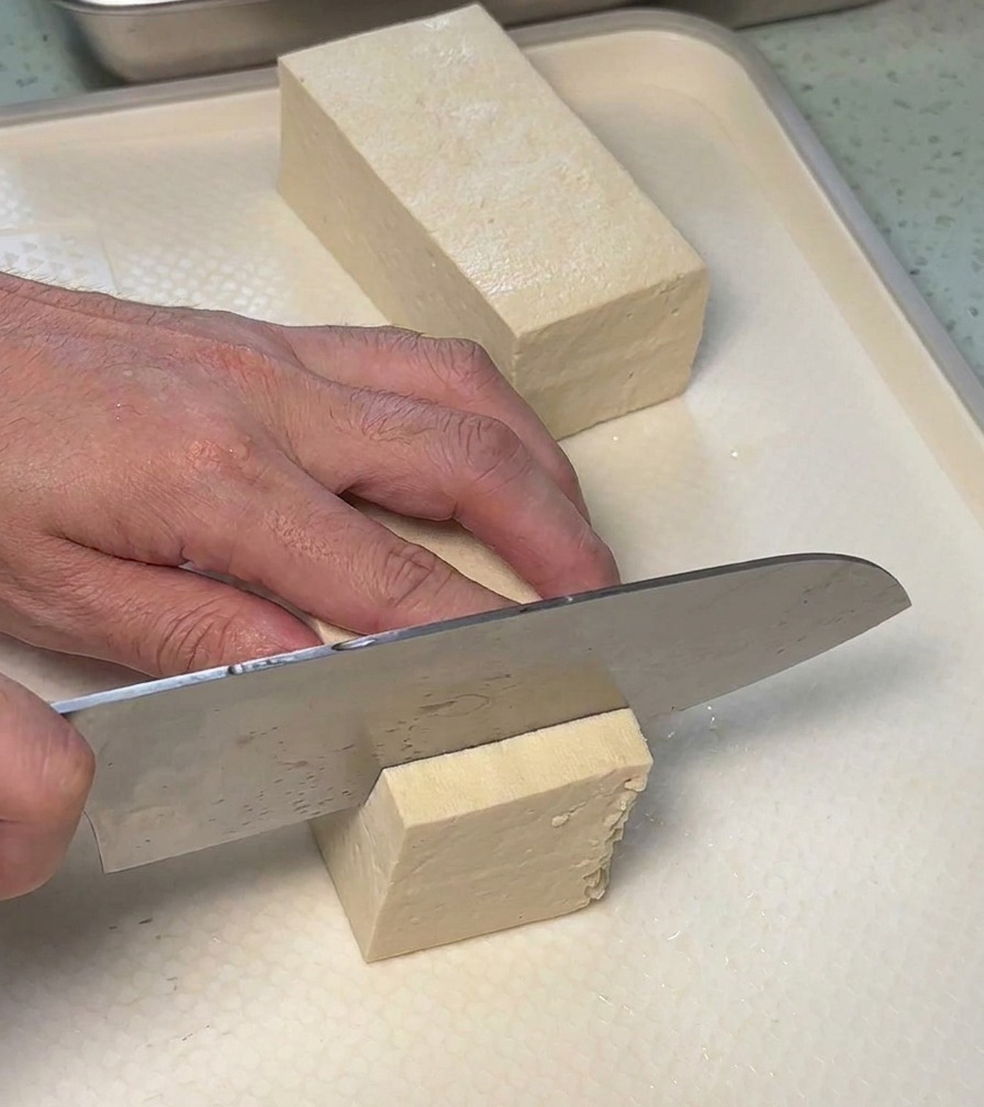 A person using a chefs knife to slice a block of firm white tofu into rectangular slabs on a white cutting board.