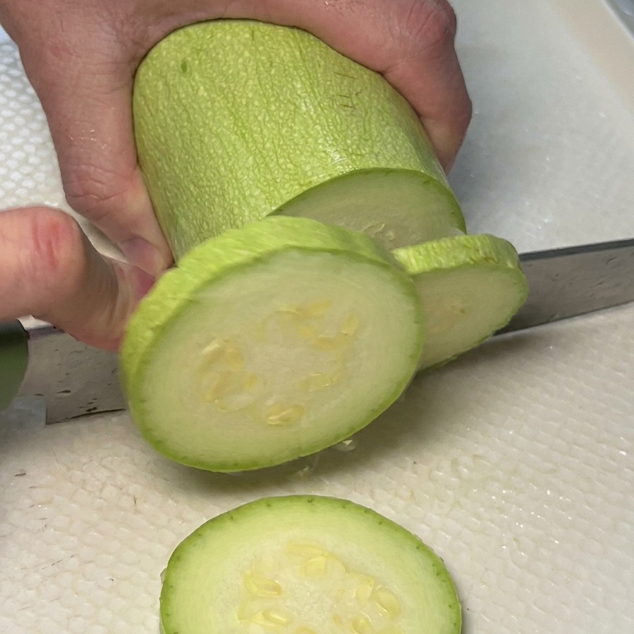 A persons hands slicing a light green zucchini into round pieces on a white cutting board.