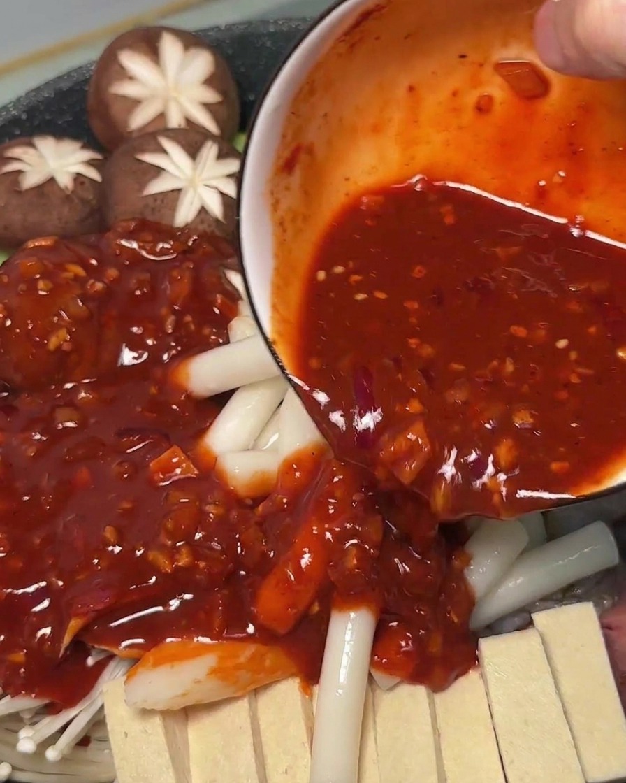 Spicy red sauce being poured from a bowl onto raw ingredients and rice cakes inside a cooking pot.