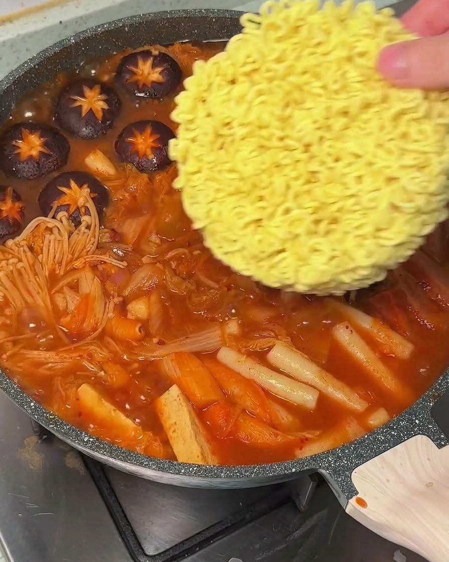 A hand placing a round block of dry instant ramen noodles into a pot of boiling spicy red soup.