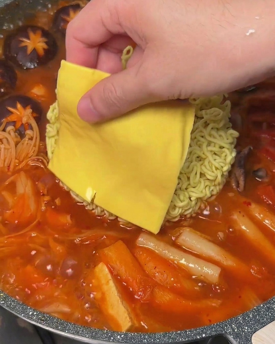 A slice of yellow cheese being placed onto a block of instant noodles in a bubbling pot of Korean Army Stew.