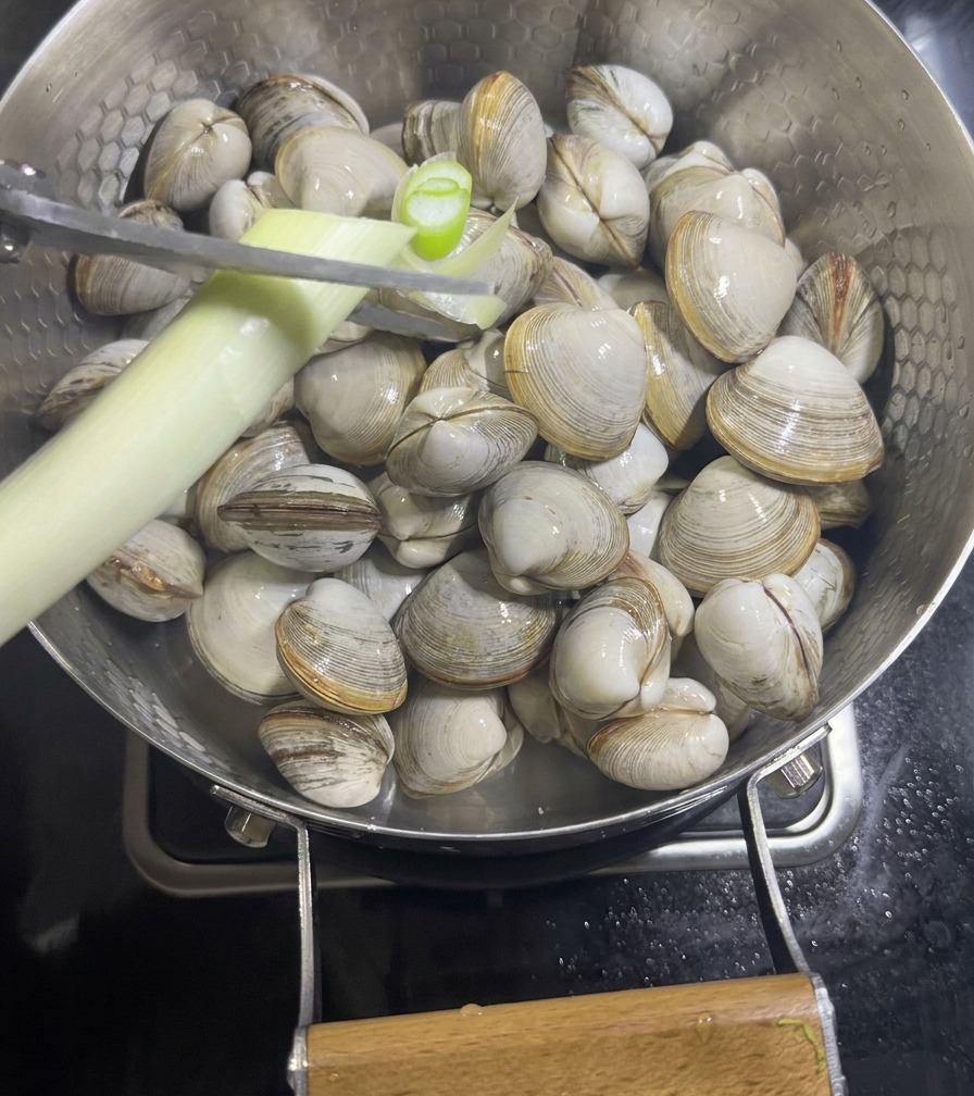 A hand using scissors to snip a stalk of green onion over a pan filled with raw white clams and sliced ginger.