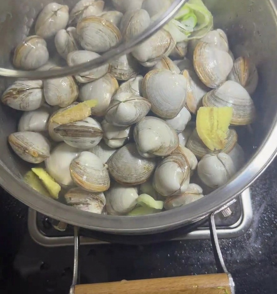 A stainless steel pan filled with white clams and aromatics being covered with a clear glass lid.