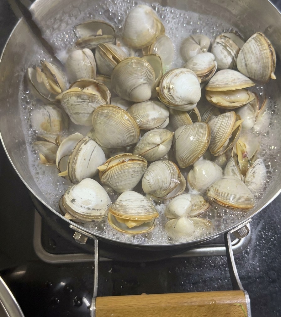 Metal tongs lifting opened, cooked white clams out of a pan with bubbling, milky seafood broth.