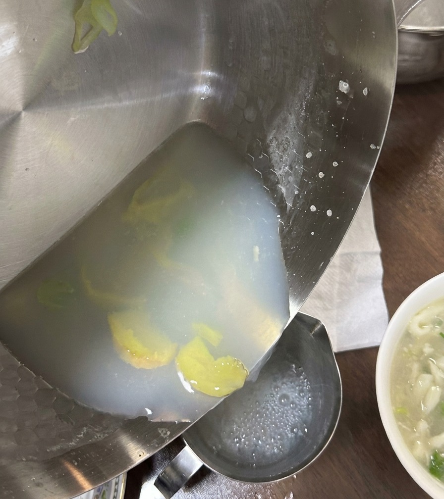 Milky white clam broth being poured from a stainless steel pan into a container, leaving ginger slices and sediment behind.