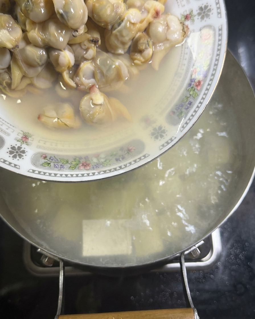 A plate of shucked, cooked clam meat is being emptied into a pot of simmering white broth that contains cubes of silken tofu.