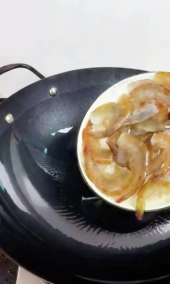 A hand holding a white plate, sliding fresh, translucent grey shrimp into a preheated black iron wok.