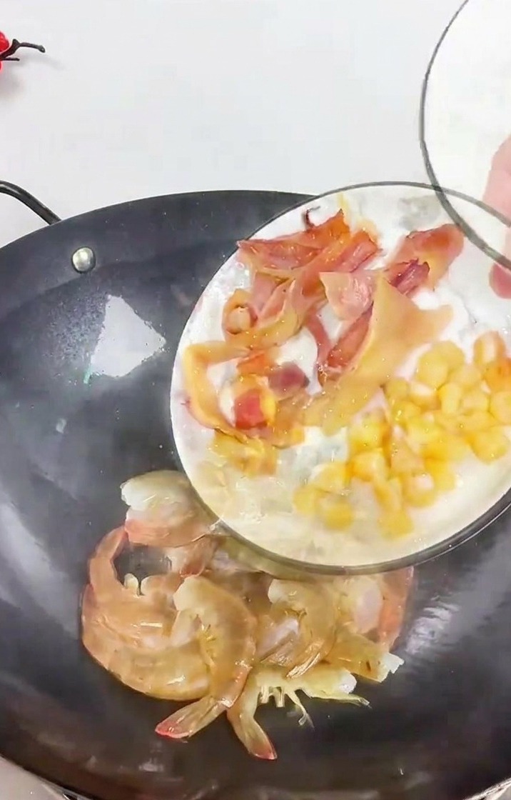 A person pouring a plate of sliced squid and dried scallops into a dark wok containing fresh whole shrimp and ginger slivers.