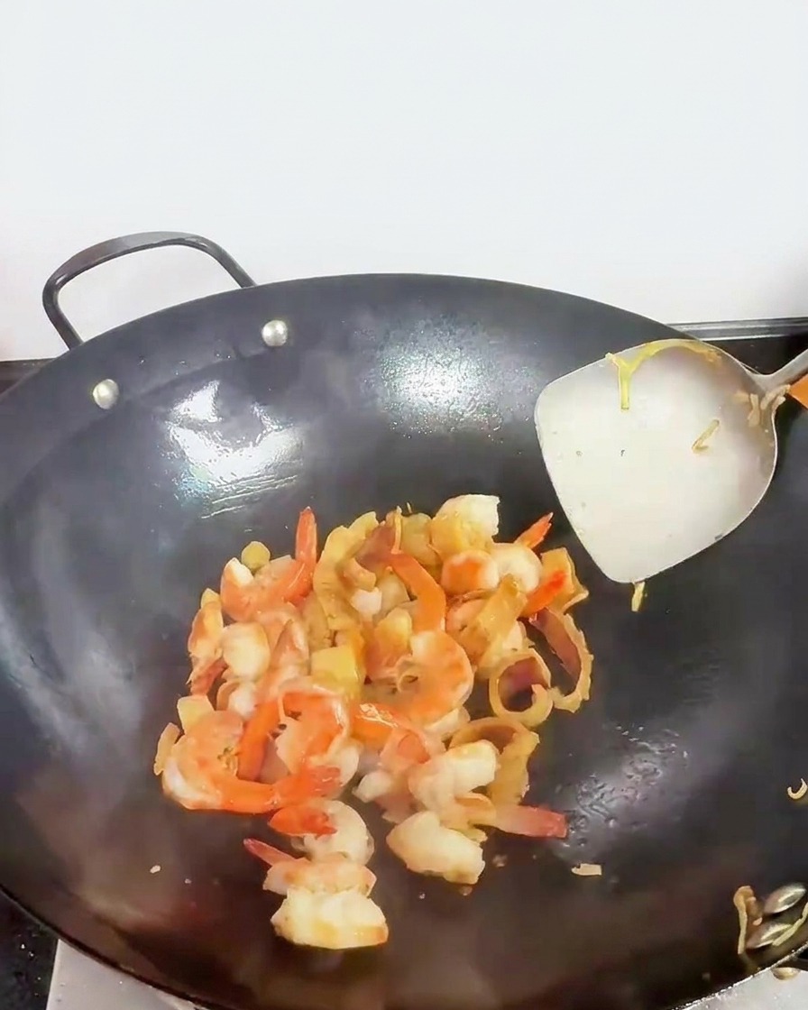 Seafood pieces, including pink-colored shrimp, being tossed in a wok with a metal spatula.