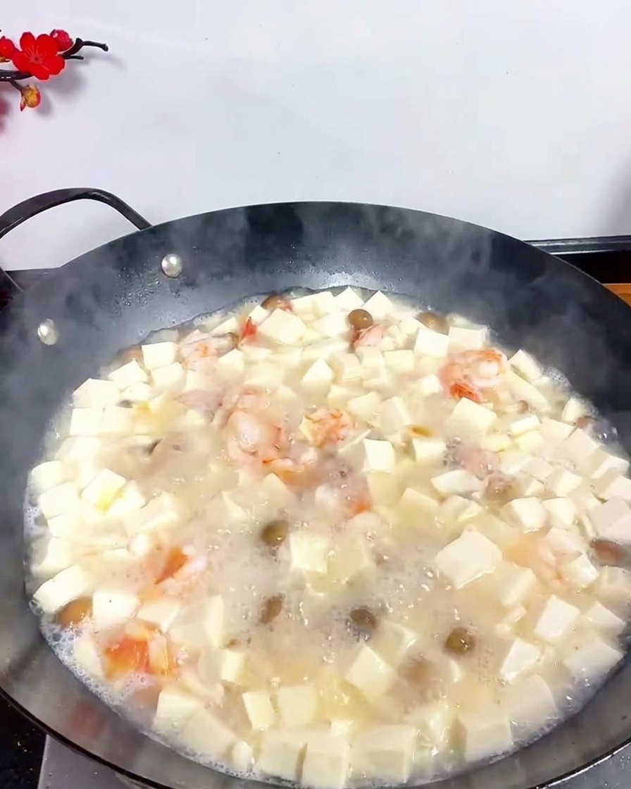A metal spoon sprinkling fine white pepper powder into a bubbling tofu and seafood soup in a black wok.
