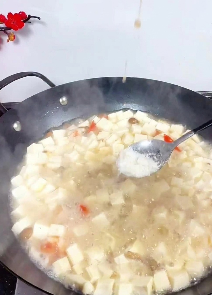 A metal spoon sprinkling white salt into a large wok filled with simmering tofu cubes and seafood in a light broth.