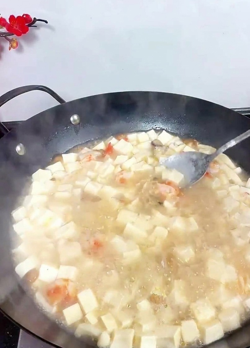 A spoon stirring a bubbling wok of white tofu soup to incorporate added seasonings.