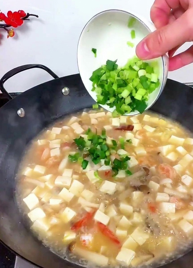 A hand tilting a small white bowl to pour bright green chopped celery and garlic sprouts into a boiling wok of tofu soup.