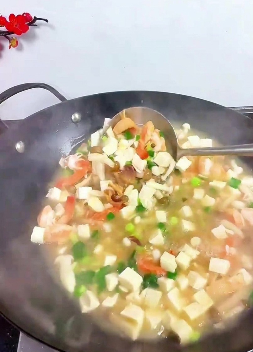 A metal ladle lifting and mixing a variety of ingredients including tofu cubes, shrimp, and green herbs in a steaming wok.