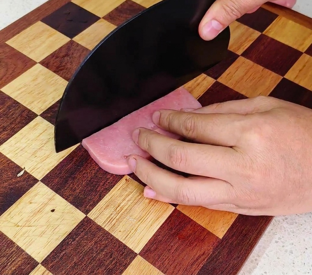 A person using a large black chefs knife to dice a pink ham sausage on a checkered wooden cutting board.