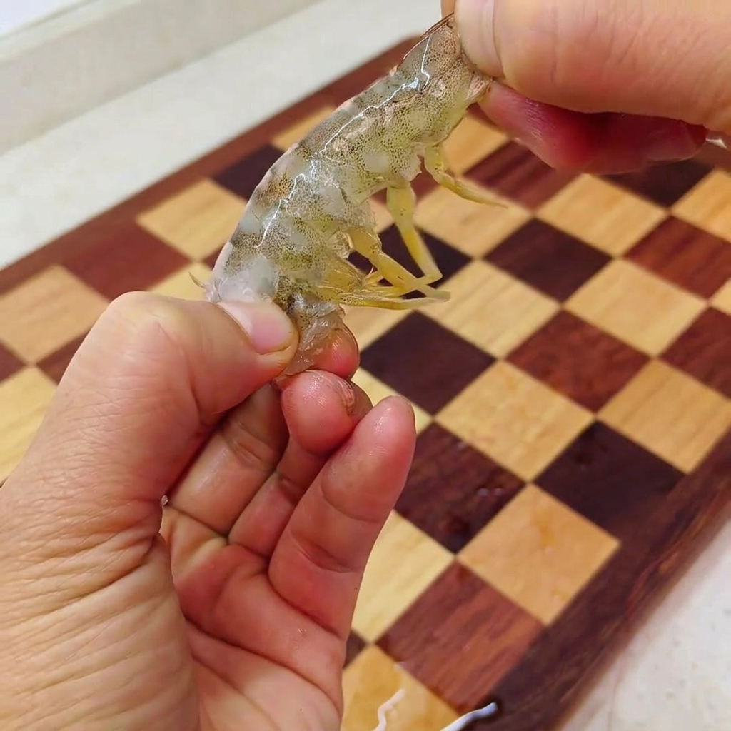 Two hands holding a partially peeled raw shrimp over a checkered wooden cutting board, pulling the meat out of the tail shell.