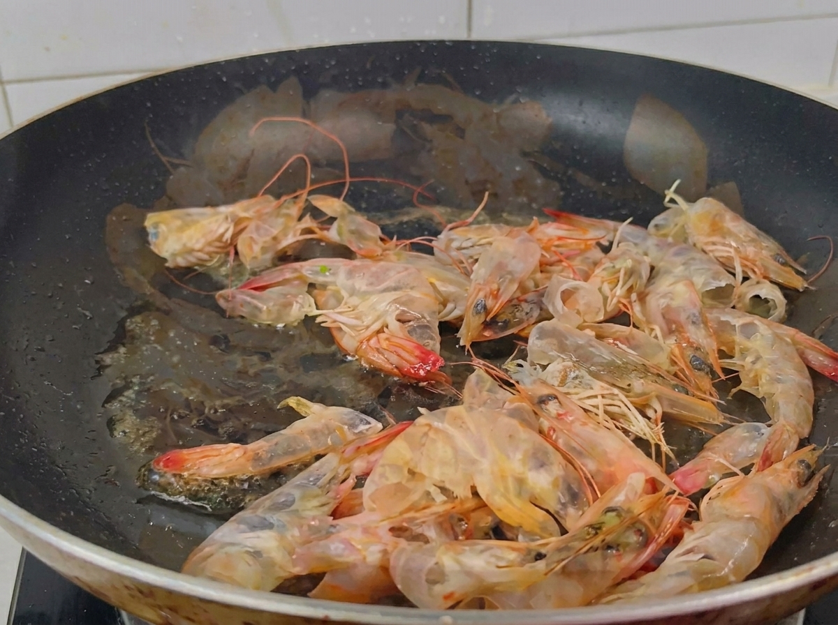 A black frying pan filled with numerous translucent and orange-tinted shrimp shells being stir-fried in shimmering oil.