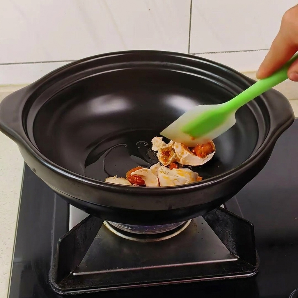 A green spatula stirring pieces of salted duck egg in a black clay pot until they become bubbly and foamy.