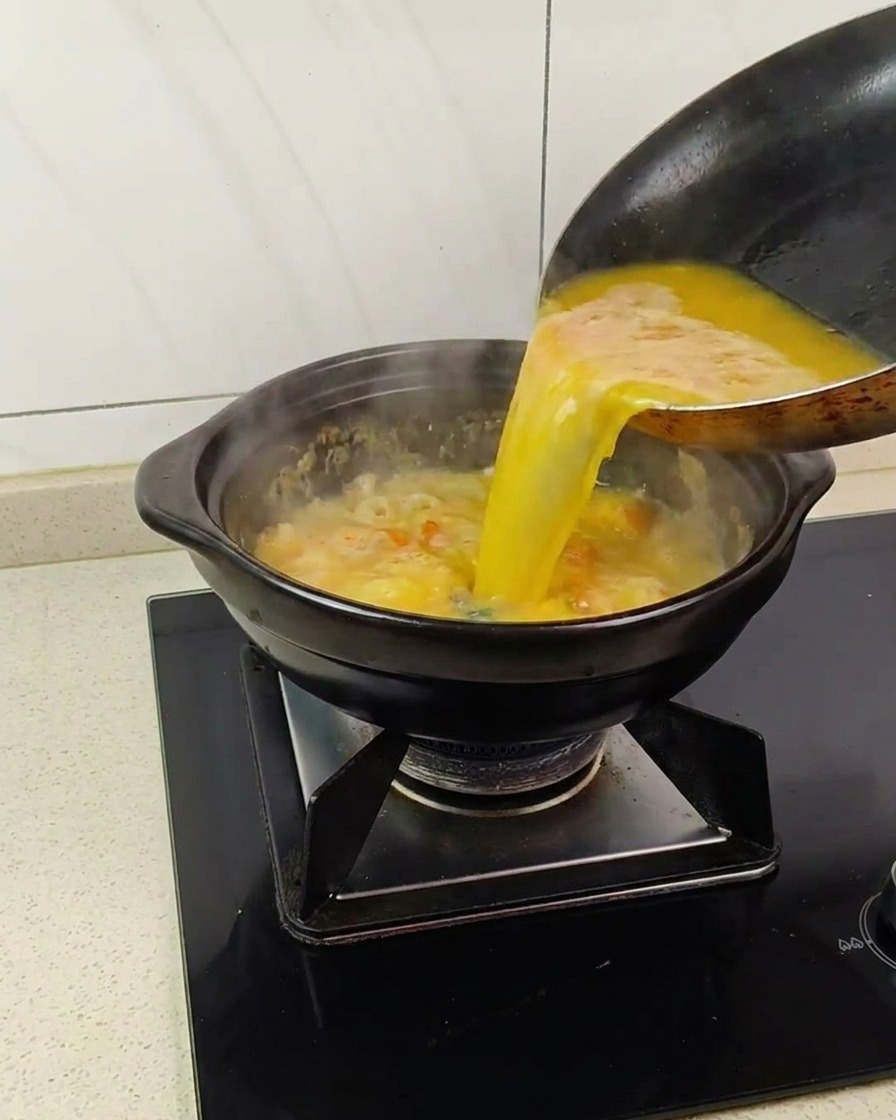 Golden-yellow liquid being poured from a pan into a black clay pot containing a bubbling, sandy egg yolk mixture.