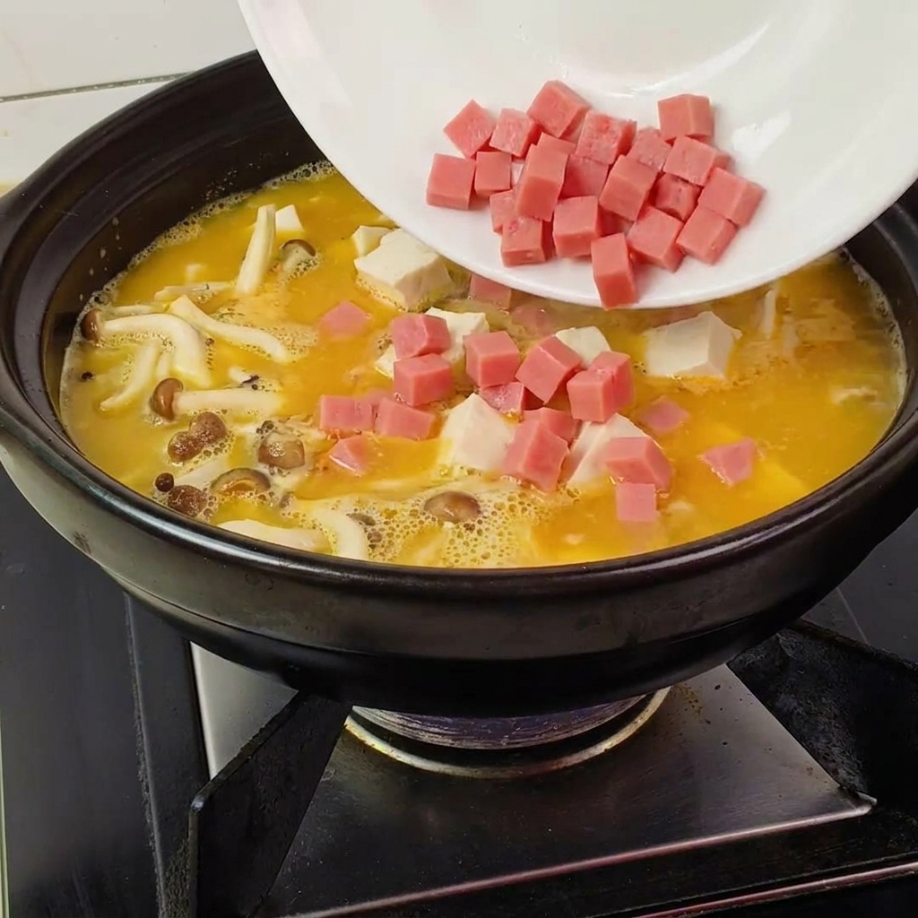 Diced pink ham being poured from a white plate into a black clay pot filled with yellow broth, white mushrooms, and tofu.