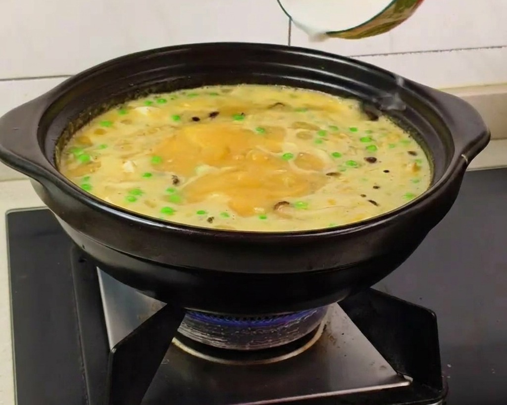 A hand pouring a white starch slurry from a small ceramic bowl into a bubbling yellow soup containing peas and mushrooms in a black clay pot.