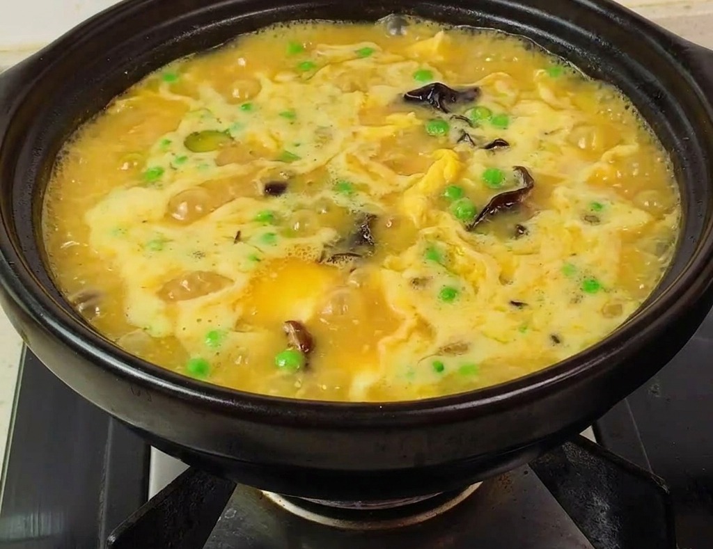 A black clay pot filled with a thick, bubbling yellow soup featuring shrimp, green peas, and mushrooms on a stove burner.