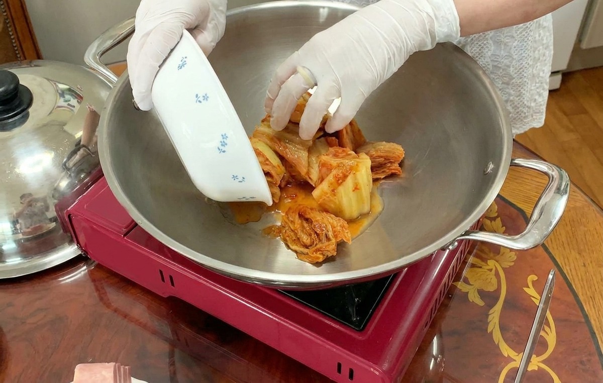 A person wearing white gloves adds sliced kimchi from a small floral bowl into the center of a large silver pot sitting on a red portable stove.