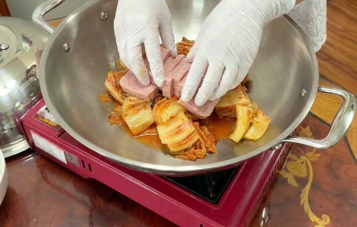 Gloved hands carefully arranging thick rectangular slices of pink ham over a bed of kimchi in a stainless steel wok.
