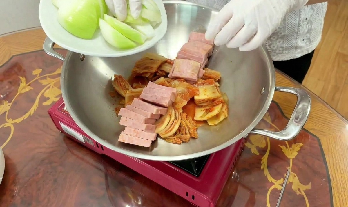 A cook adds fresh white onion wedges from a plate into a cooking pot already filled with kimchi and sliced spam.