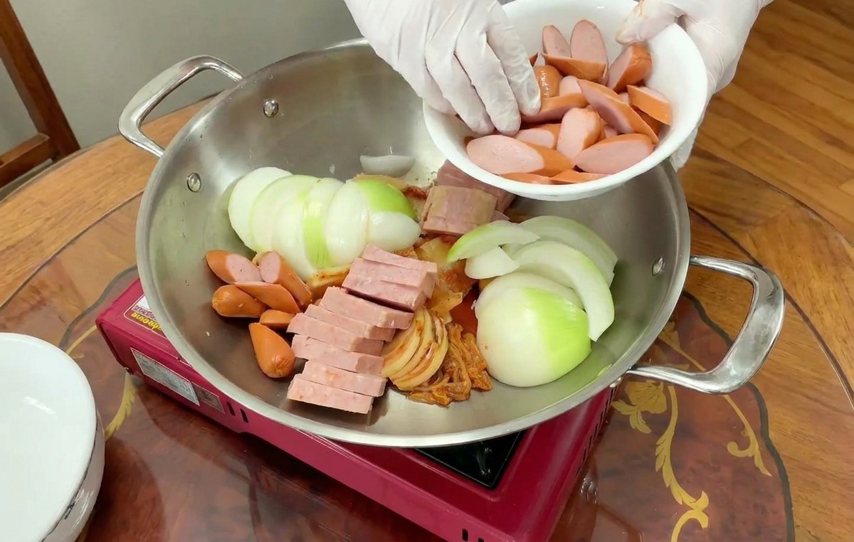 Sliced red-skinned sausages being poured from a white bowl into a pot containing onions, ham, and kimchi.