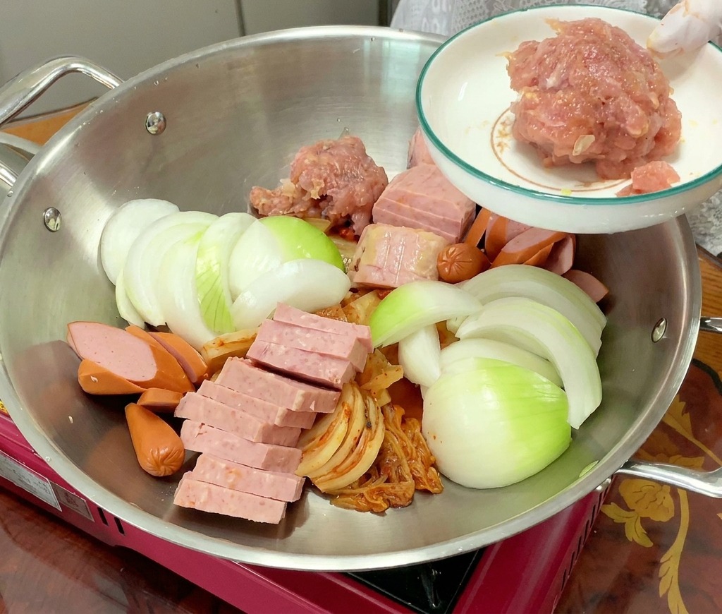 A mound of pink, seasoned ground pork is added to a pot filled with arranged kimchi, spam, sausages, and onions.