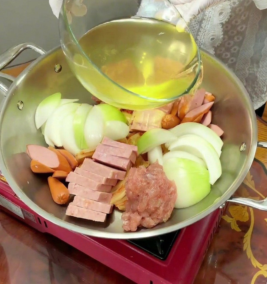 Clear yellow liquid being poured from a glass bowl into a stainless steel pot filled with raw stew ingredients.
